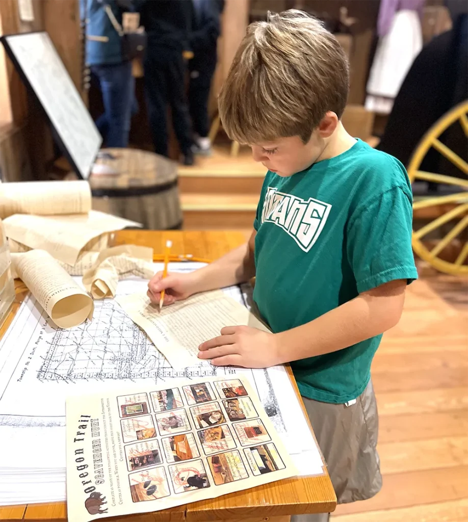 elementary aged boy with a tshirt and shorts writing down notes about the Oregon Trail on a field trip