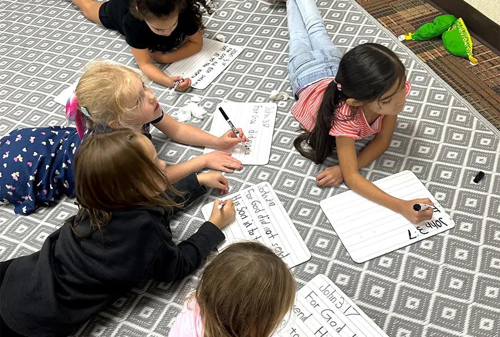 five elementary girls practicing writing while laying on their stomachs on the floor at Riviera Christian School